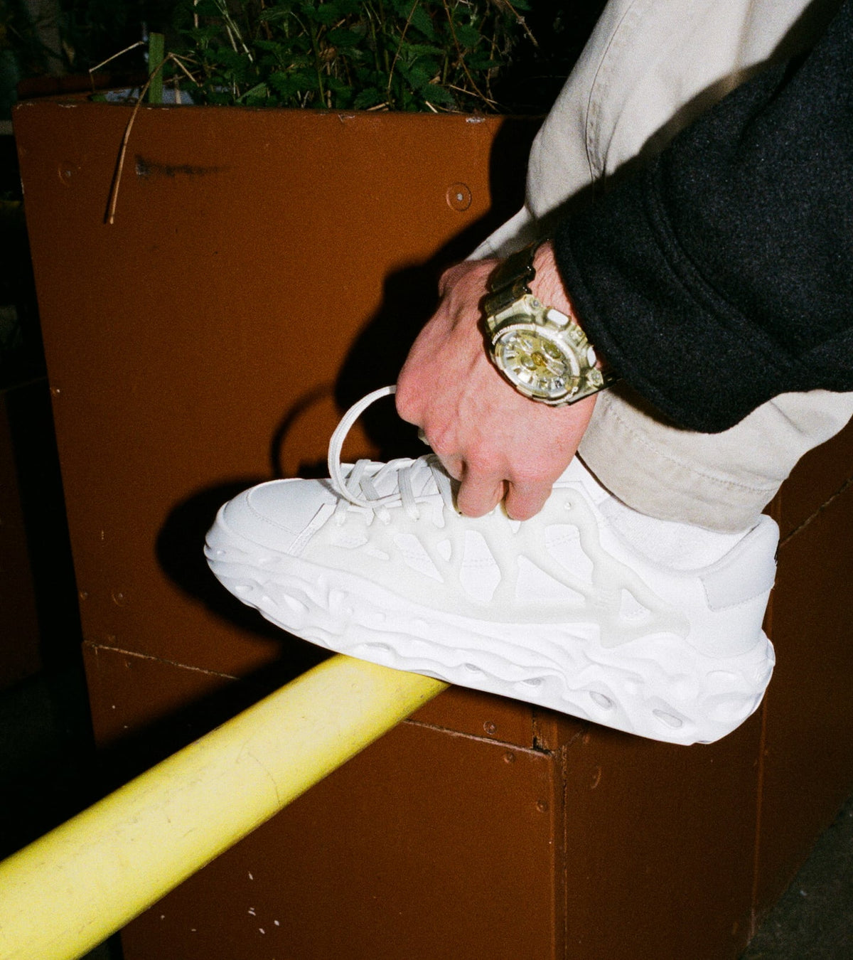 Close-up of the left FFS RADICLE LOW OG shoe from the lateral view, worn by a person who is standing with it on a yellow bar and tying it