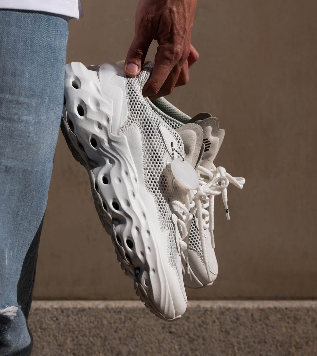 A pair of the Seed.One in the Blanc Canvas colorway from the lateral view held by a hand next to a person's leg in blue jeans