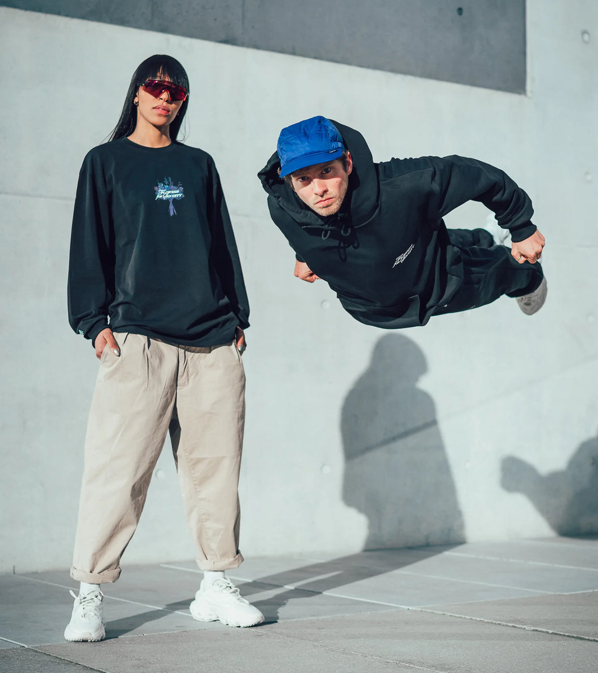 Two models posing in apparel of the Bouquet Collection in front of a concrete building, the left person is standing while the right seems to be standing horizontally in the air